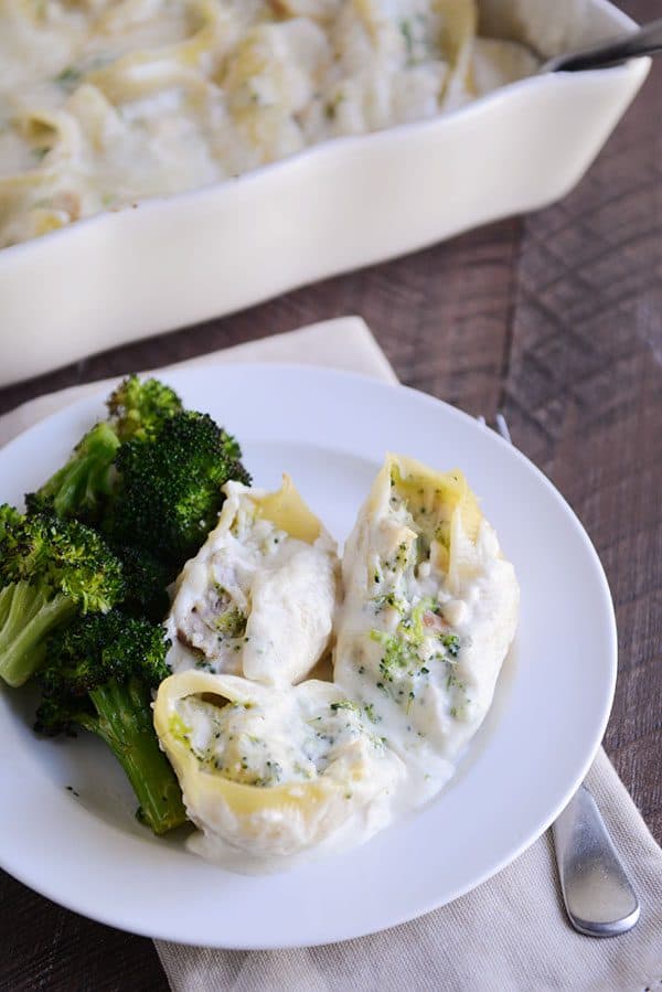 Un plato blanco con tres conchas gigantes rellenas de pasta Alfredo, junto a un brócoli cocido.