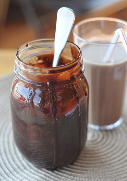 Un tarro de albañil de jarabe de chocolate con agua que gotea al lado y una cuchara, colocado frente a un vaso de leche con chocolate.