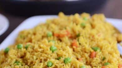 Un plato blanco se llena de arroz con guisantes y zanahorias, con una olla instantánea de fondo.