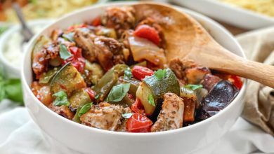 Cuenco blanco y cuchara de madera con pollo, calabacín, tomates y otras verduras.