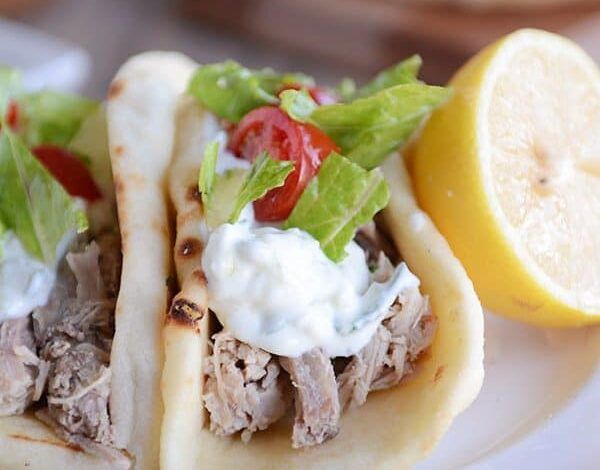 En un plato blanco había dos tacos de cerdo griegos con lechuga y tomates encima y medio limón a un lado.