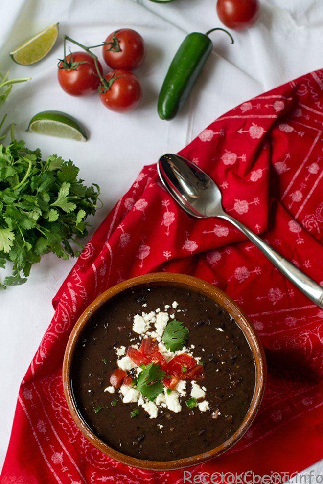 Sopa de frijoles negros en un tazón cubierto con queso fresco, tomates y perejil. Sobre una tela estampada roja, hay cucharas y varios ingredientes frescos alrededor.