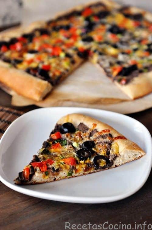 Se coloca un trozo de pizza de frijoles negros y verduras en un plato blanco, y el resto de la pizza se coloca detrás del plato.