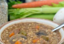 Sopa de carne y cebada con zanahorias y champiñones en un cuenco blanco y marrón con una cuchara.