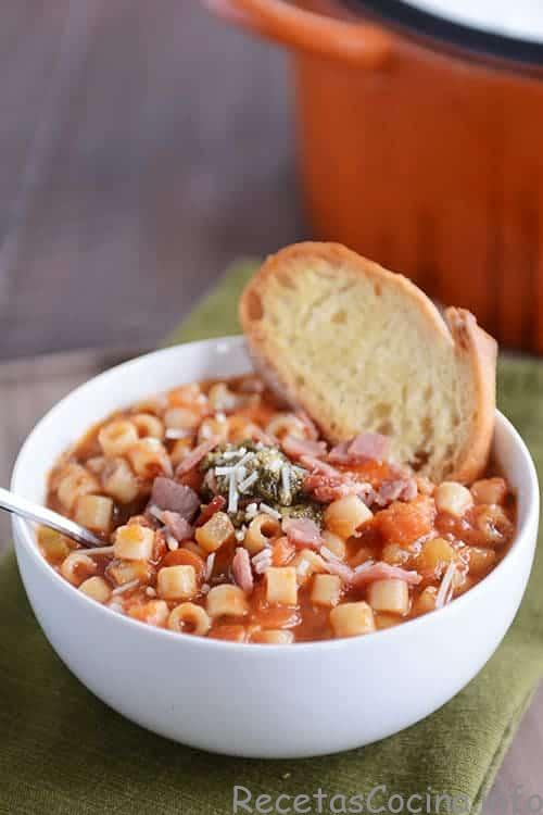 Un cuenco blanco lleno de sopa minestrone italiana de invierno, con un trozo de pan de ajo al lado.