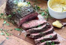 London broil on a wooden board with several slices cut, topped with butter.