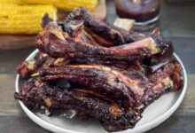 BBQ grilled beef ribs stacked on a plate with BBQ sauce and corn on the cob in the background.